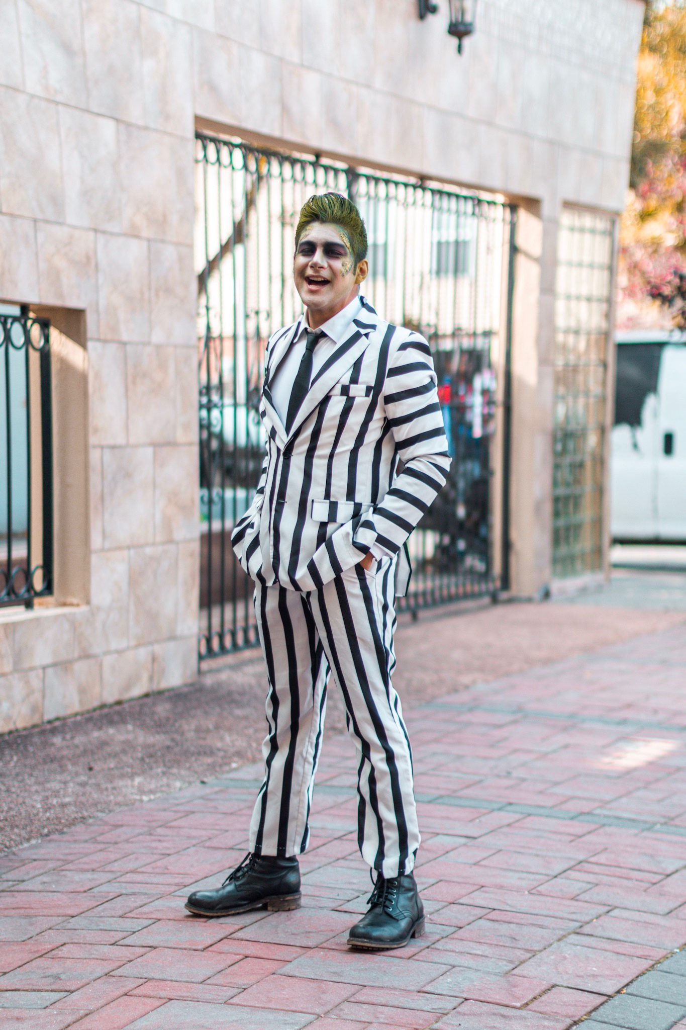In the image, there's a person standing on a brick pavement with a cheerful demeanor. They are dressed in a unique, bold suit that has black and white vertical stripes, evoking the style of a classic movie character known for his mischievous and whimsical personality. The suit is well-fitted, featuring a blazer and trousers that match in pattern. The person's hair is styled upwards and colored a vibrant green, complementing the theatrical theme of the outfit. Their face is painted white with darkened eyes and a wide, exaggerated smile, adding to the playful and dramatic effect. Behind them is a background of a gated fence and a building with a stone facade, which brings a sense of urbanity to the scene. Their pose and expression suggest confidence and a sense of fun, embodying the spirit of a character who is both entertaining and a bit unconventional.

| BEETLEJUICE cosplay | BEETLEJUICE costume | striped suit | BEETLEJUICE | BEETLEJUICE suit | dandy in the bronx