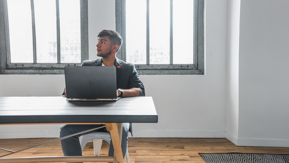 “Man in a blazer sitting at a table with a laptop, looking to the side, in a well-lit room with large windows.”