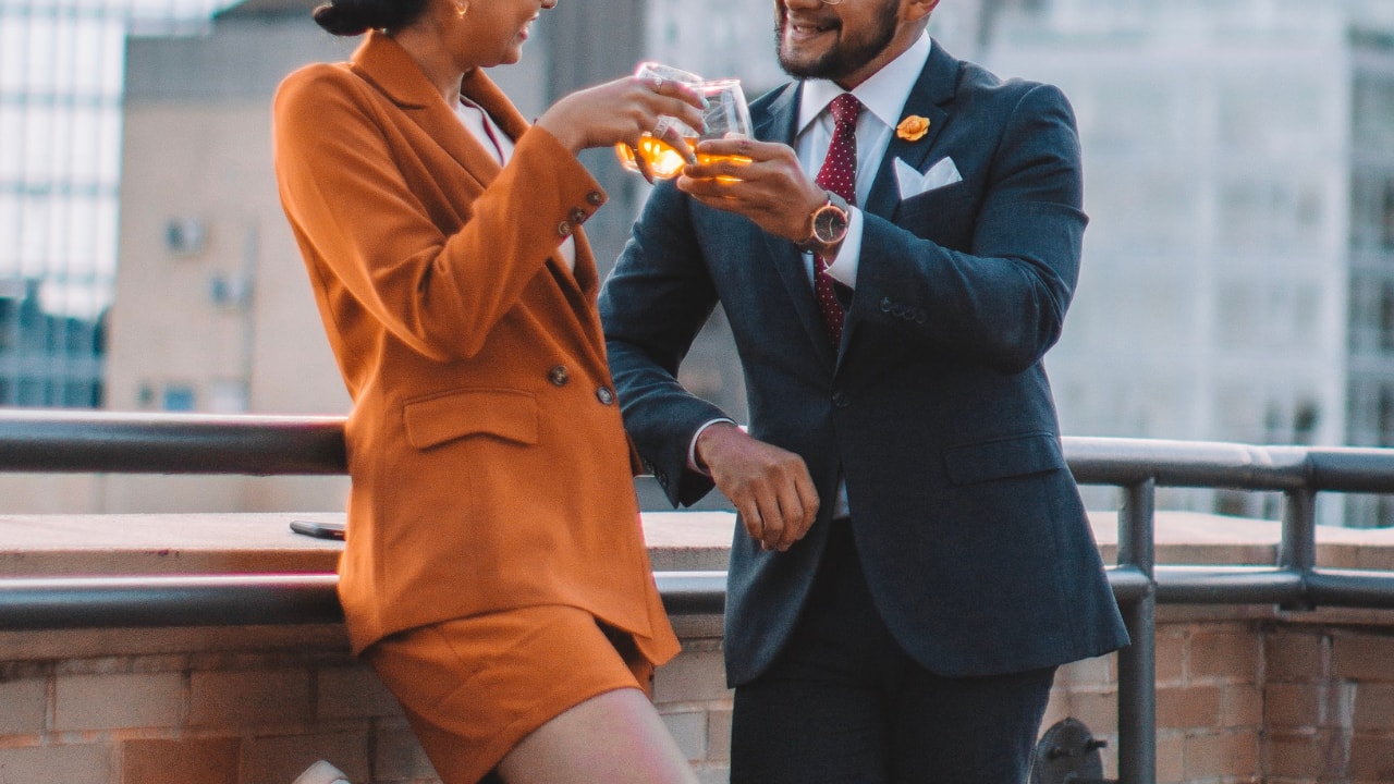 woman in an orange suit - man in navy suit - people toasting their drinks