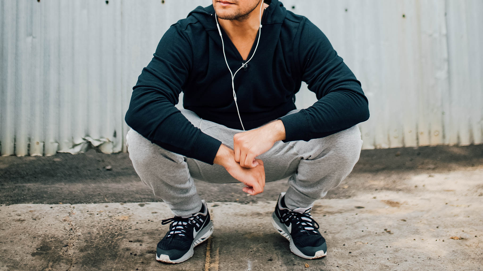 man in gym clothes squatting - man in running clothes - man wearing grey joggers - grey sweat pants - black hoodie